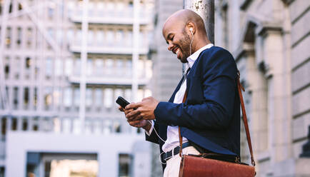 Fröhlicher junger Geschäftsmann mit einem Smartphone, der in einer Straße in der Stadt spazieren geht. Afrikanischer Mann im Anzug mit Kopfhörern, der im Freien Musik auf einem Mobiltelefon hört. - JLPSF19818