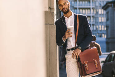 Porträt eines gut aussehenden jungen Mannes, der sich mit einem Kaffee im Freien an die Wand lehnt. Afrikanischer Geschäftsmann mit Handtasche, der Kopfhörer trägt und eine Tasse Kaffee in der Hand hält. - JLPSF19819