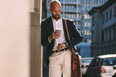 Junger afrikanischer Geschäftsmann, der sich im Freien an eine Wand lehnt und die Zeit auf seiner Armbanduhr überprüft. Mann im Anzug, der draußen in der Stadt auf jemanden wartet. - JLPSF19820