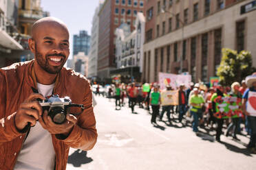 Ein lächelnder afrikanischer Fotograf fotografiert Aktivisten auf der Straße. Ein Mann fotografiert Menschen, die an einer Kundgebung auf einer Straße in der Stadt teilnehmen. - JLPSF19826