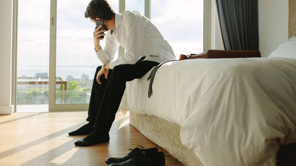 Ein Geschäftsmann sitzt auf seinem Bett und spricht mit seinem Handy. Ein Mann telefoniert, während er sich auf die Arbeit vorbereitet. - JLPSF19831