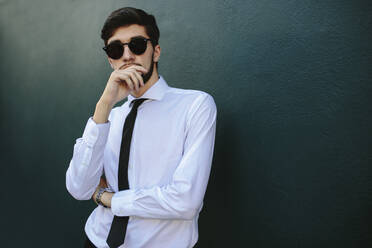 Ein elegant gekleideter junger Geschäftsmann steht an einer dunklen Wand, ein Mann in formeller Kleidung posiert mit Sonnenbrille. - JLPSF19839