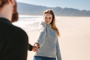 Lächelnde Frau, die die Hand eines Mannes hält, während sie am Strand spazieren geht. Glückliches junges Paar, das am Meer spazieren geht. - JLPSF19843