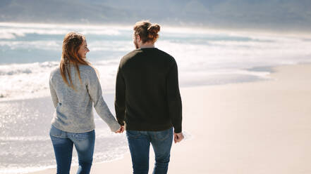 Rückansicht eines romantischen Paares, das Hand in Hand am Strand spazieren geht. Mann und Frau machen einen Spaziergang am Meer. - JLPSF19847