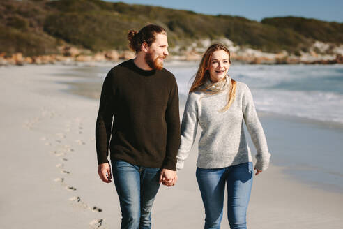 Ein lächelnder Mann und eine lächelnde Frau gehen zusammen am Meer spazieren und halten sich an den Händen. - JLPSF19848
