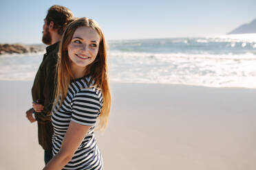 Hübsche junge Frau schaut zurück und lächelt, während sie mit ihrem Freund am Strand spazieren geht. Junges Paar am Meer. - JLPSF19860