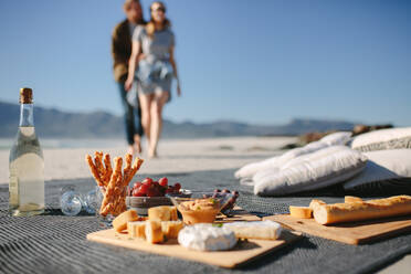 Essen und Trinken auf einer Decke am Strand mit einem Paar, das darauf zugeht. Picknick am Strand mit Mann und Frau im Hintergrund. - JLPSF19873