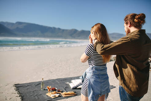 Rückansicht eines Mannes, der die Augen seiner Freundin am Strand bedeckt, um sie mit Essen und Getränken auf einer Decke zu überraschen. Der Freund bedeckt die Augen seiner Frau, um sie zu überraschen. - JLPSF19874