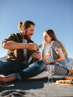 Glücklicher junger Mann öffnet eine Flasche Champagner am Strand mit seiner Freundin, die auf einer Decke sitzt. Junges Paar beim Picknick am Meer öffnet eine Champagnerflasche. - JLPSF19875