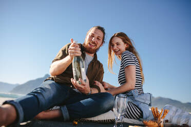 Beautiful couple on picnic at the sea shore opening a champagne bottle. Man and woman having picnic at the beach with food and drinks. - JLPSF19876