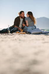 Beautiful couple enjoying in a good mood and picnic day on the beach. Low angle view of man and woman sitting by the beach with food and drinks. - JLPSF19878