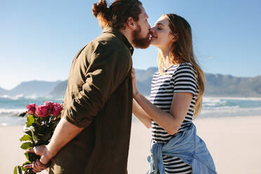 Mann hält Blumen hinter sich und küsst seine Freundin am Strand. Verliebtes Paar küsst sich am Strand. - JLPSF19881