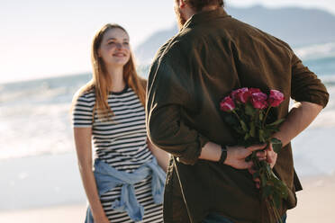 Junges verliebtes Paar bei einem romantischen Rendezvous. Mann hält Überraschungsstrauß von Rosen für schöne junge Frau am Strand. - JLPSF19886