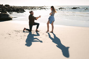 Mann kniet und macht Frau am Meer einen Heiratsantrag. Mann macht Frau am Strand mit einem imaginären Ring einen Heiratsantrag. - JLPSF19892