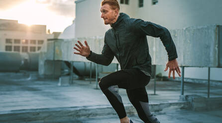 Seitenansicht eines Sportlers, der auf dem Dach eines Gebäudes mit Sonnenschein im Hintergrund läuft. Männlicher Sportler beim Fitnesstraining auf der Terrasse eines Gebäudes bei Sonnenaufgang. - JLPSF19896