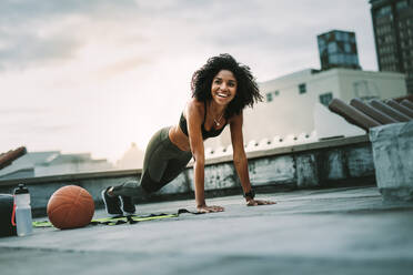 Lächelnde Sportlerin macht Liegestütze auf dem Dach mit Gymnastikbällen an ihrer Seite. Fröhliche Frau in Fitnesskleidung beim Training auf der Terrasse. - JLPSF19898