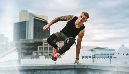 Man doing fitness training on rooftop. Muscular man jumping on to the terrace from the rooftop fence with one hand on fence. - JLPSF19901