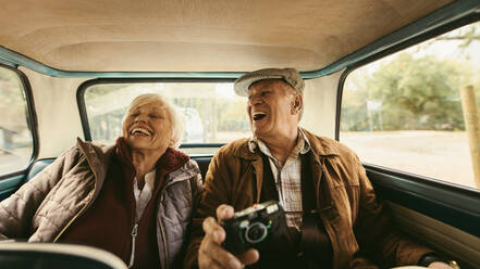 Fröhliches altes Paar, das auf dem Rücksitz seines Autos sitzt und lacht. Älterer Mann und Frau genießen es, zusammen in einem alten Auto zu reisen. - JLPSF19905