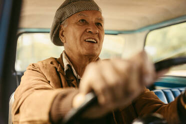 Lächelnder älterer Mann, der ein altes Auto fährt. Glücklicher alter Mann mit Mütze, der sich am Fahren seines Autos erfreut. - JLPSF19909
