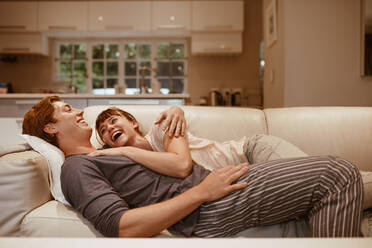 Glückliches Paar, das sich unterhält und lacht, während es zu Hause auf der Couch liegt. Die Frau schläft auf dem Arm ihres Mannes, der auf der Couch schläft und sich unterhält. - JLPSF19915