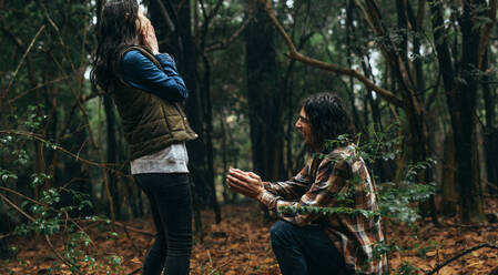 Couple in forest, with man proposing his girlfriend. Man kneeling holding a ring and making a marriage proposal to woman with surprised expression. - JLPSF19927