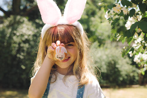 Close up of a smiling girl holding a painted easter egg in front of her face. Girl wearing rabbit ear headband having fun playing with painted eater egg. - JLPSF19935