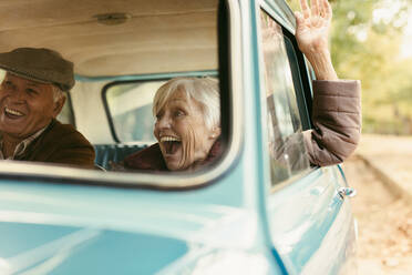 Senior woman enjoying road trip with her husband. Excited old female raising her hand out of the car and laughing. Senior couple enjoying on their road trip. - JLPSF19942