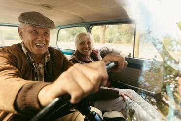 Smiling senior man driving a old car with a woman sitting next to him in passenger seat. Happy senior couple going on a road trip. - JLPSF19944