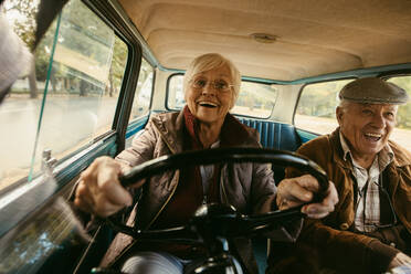 Smiling elderly woman in warm clothing driving car with husband sitting in passenger seat. Old couple having a great time on their drive. - JLPSF19946