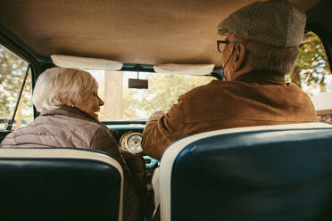 Rear view of old couple driving a car. Senior man wearing hat driving the car and talking to old woman sitting on passenger seat. - JLPSF19947