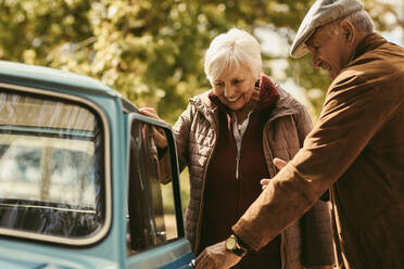 Fürsorglicher Herr mit warmem Mantel und Hut, der das Auto für seine Frau öffnet, während sie zusammen unterwegs sind. Älteres Paar, das sich zu einem Road Trip verabredet. - JLPSF19948