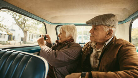Senior man sitting in backseat of car with wife taking pictures with digital camera. Old couple taking photographs on road trip. - JLPSF19950