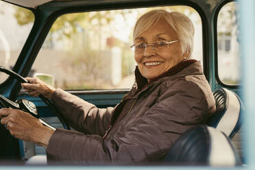 Porträt einer schönen älteren Frau in warmer Kleidung, die ein altes Auto fährt. Lächelnde ältere Frau sitzt auf dem Fahrersitz eines Autos und schaut in die Kamera. - JLPSF19953