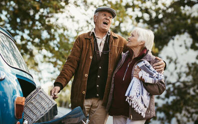 Älterer Mann, der den Picknickkorb aus dem Kofferraum holt und einen Ausdruck macht, da er sehr schwer ist, und seine Frau hält. Älteres Paar, das an einem Wintertag ein Picknick macht. - JLPSF19957