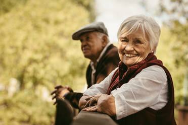 Portrait of beautiful senior woman standing by a wooden railing with her partner at the back at park. Smiling old woman at park with her husband. - JLPSF19963