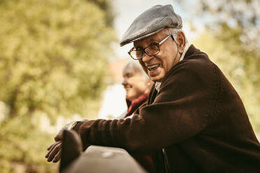 Portrait of happy senior man wearing glasses and cap standing by a wooden railing with a woman at the back in park. Smiling old man at park on a winter day. - JLPSF19964
