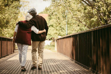 Rückansicht eines älteren Mannes und einer älteren Frau, die an einem Wintertag durch einen Park spazieren gehen und sich in den Armen liegen. Älteres Paar in warmer Kleidung, das gemeinsam im Park spazieren geht. - JLPSF19967