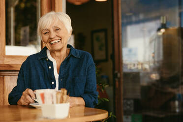 Porträt einer lächelnden alten Frau im Café mit einer Tasse Kaffee auf dem Tisch. Schöne ältere Frau im Café schaut in die Kamera und lächelt. - JLPSF19968