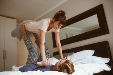 Happy couple playing in bed and having fun. Man lying on bed and lifting his wife with his feet holding her hands. - JLPSF19982