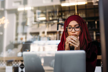 Muslimische Frau mit Hijab, die in einem Café sitzt und Kaffee trinkt. Islamische Frau, die in einem Café einen Morgenkaffee trinkt und einen Laptop benutzt. - JLPSF19992