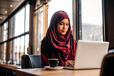 Pretty islamic female sitting at cafe table and working on laptop with coffee in side. Businesswoman in hijab working from a coffee shop. - JLPSF20007