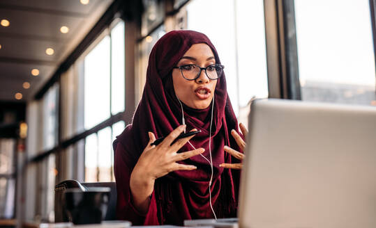 Geschäftsfrau in Hijab, die einen Video-Chat auf einem Laptop führt, während sie in einem Café sitzt. Frau sitzt in einem Café und führt einen Videoanruf mit Kopfhörern und Laptop durch. - JLPSF20011