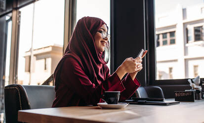 Woman in hijab sitting at cafe using mobile phone. Islamic woman social networking on her smartphone while sitting at coffee shop. - JLPSF20013