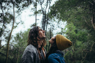 Junges Paar im Wald, das lächelnd nach oben schaut. Junger Mann steht mit seiner Freundin im Wald, beide schauen nach oben und lächeln. - JLPSF20031