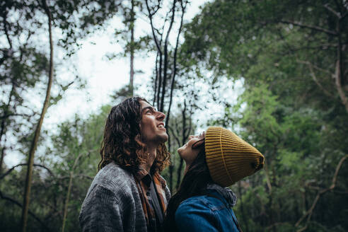 Young couple looking up and smiling in forest. Young man standing with his girlfriend in a forest, both looking up and smiling. - JLPSF20031