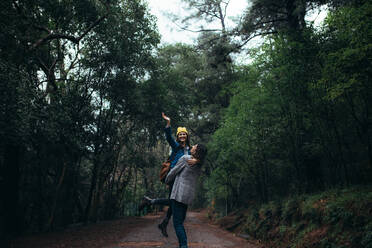 Man carrying her girlfriend raising hand and smiling. Couple enjoying themselves at forest on rainy day. - JLPSF20040