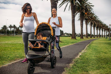 Zwei Frauen gehen mit einem Kinderwagen in einem Park spazieren. Schwangere Frau auf einem Morgenspaziergang mit ihrem Baby in einem Kinderwagen. - JLPSF20055