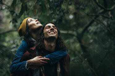 Glücklicher junger Mann trägt seine Frau auf dem Rücken im Wald und genießt den Regen. Hübsches Paar, das im Park bei Regen Huckepack nimmt. - JLPSF20061