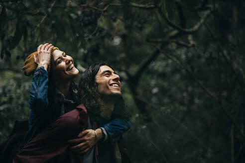 Young couple at forest piggybacking under the rain. Man carrying his smiling girlfriend on back at the forest on a rainy day. - JLPSF20062