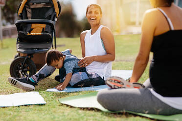 Smiling mother with her kid sitting in park on a yoga mat with a baby stroller beside her. Women in park with kid doing yoga early in the morning. - JLPSF20065
